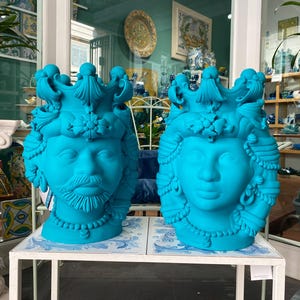 May include: Two turquoise ceramic planter heads with ornate details. The heads are facing forward and have a crown-like design on top. The planters are on a white table.