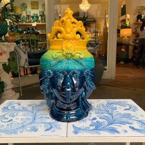 May include: A blue and yellow ceramic head vase with a crown on top. The vase is decorated with a floral pattern and has a blue and white tiled surface underneath.