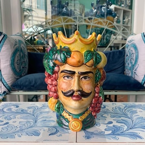 May include: A ceramic head vase with a crown, mustache, and fruit details. The face is painted with realistic features, and the crown is yellow. The vase is decorated with grapes, oranges, and green leaves. The vase is on a blue and white patterned surface.