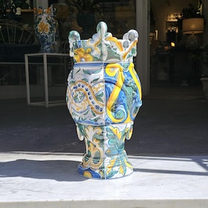 May include: A large, decorative ceramic vase with a blue, green, yellow, and white floral pattern. The vase has a square base and a rounded top.