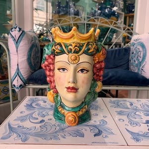 May include: A ceramic vase shaped like a woman's head with a crown, grapes, and floral details. The vase is painted in shades of green, yellow, red, and white.