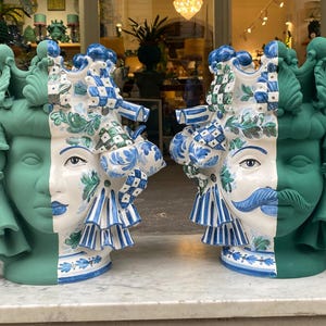 May include: Two ceramic planters shaped like human heads with green and blue painted details. The planters are split in half, with one side painted in a green glaze and the other side painted in a blue and white pattern.