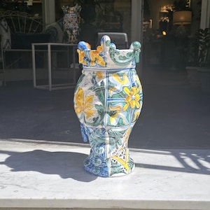 May include: A large, ceramic vase with a crown-like top and a colorful, geometric pattern. The vase is decorated with blue, green, yellow, and white floral motifs.