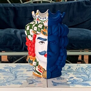 May include: A ceramic vase with a unique design featuring two faces, one with a green and white checkered hat and the other with a blue crown. The vase is painted in vibrant colors, including red, blue, white, and green.