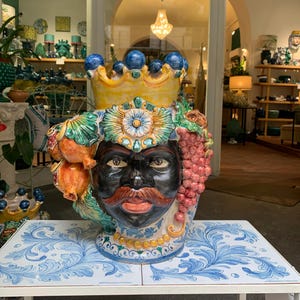 May include: A colorful ceramic vase with a human face, a crown, and fruit decorations. The vase is painted in shades of green, yellow, red, and blue.