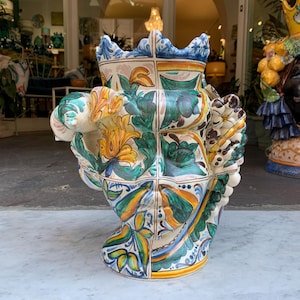 May include: A ceramic vase with a whimsical design featuring a face with floral patterns in shades of blue, green, yellow, and white.