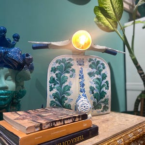 May include: A white ceramic scooter lamp with blue and green floral designs. The lamp is sitting on a stack of three books with Italian titles. The books are on a table with a gold and brown patterned surface.