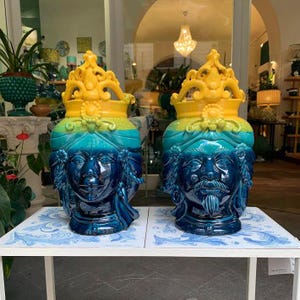 May include: Two blue and yellow ceramic vases with faces and crowns. The vases are on a white table with a blue and white tiled surface.
