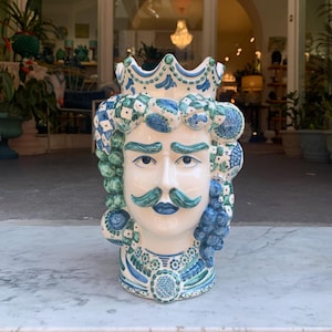 May include: A white ceramic vase shaped like a man's head with a crown, blue and green painted details, and a mustache.