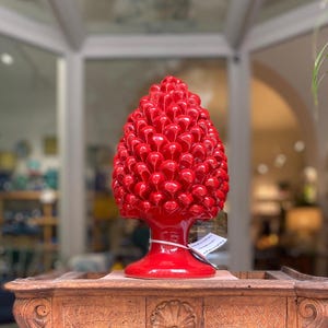 Può includere: Una scultura decorativa a forma di pigna in ceramica rosso vivo. Il pezzo presenta una superficie strutturata e dettagliata con una finitura lucida. È esposta su un tavolo di legno.