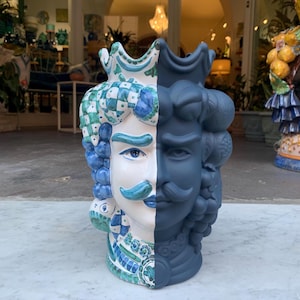 May include: A ceramic vase in the shape of a face, half painted white with blue and green details, and half painted dark blue. The vase has a crown on top and a mustache.