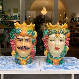 May include: Two ceramic vases shaped like human heads, one male and one female, with crowns and fruit decorations. The vases are painted in bright colors, including yellow, green, red, and orange.