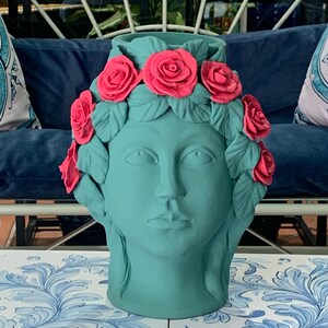May include: A teal blue ceramic vase shaped like a woman's head with a pink rose crown. The vase is a decorative home decor item.