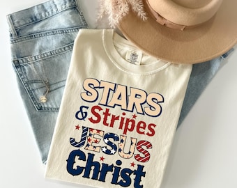 Puede incluir: Camiseta color crema con las palabras "Stars & Stripes Jesus Christ" en rojo, blanco y azul. La camiseta se combina con unos vaqueros de color azul claro, un collar dorado y un sombrero color canela. También hay una ramita de flores secas.