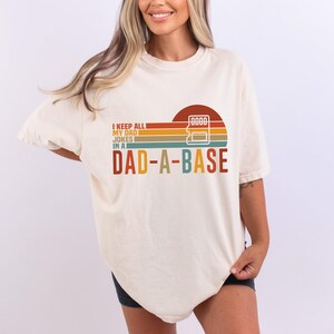 May include: Cream-colored t-shirt with a retro design. The graphic features a sunset with a calculator and the text "I KEEP ALL MY DAD JOKES IN A DAD-A-BASE" in various colors. A fun, novelty tee.