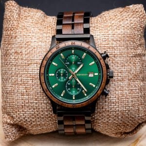 A beautiful wooden watch, with green face surrounded by a walnut bezel accompanied by a walnut and black stainless steel strap. On a display cushion on a round of wood.