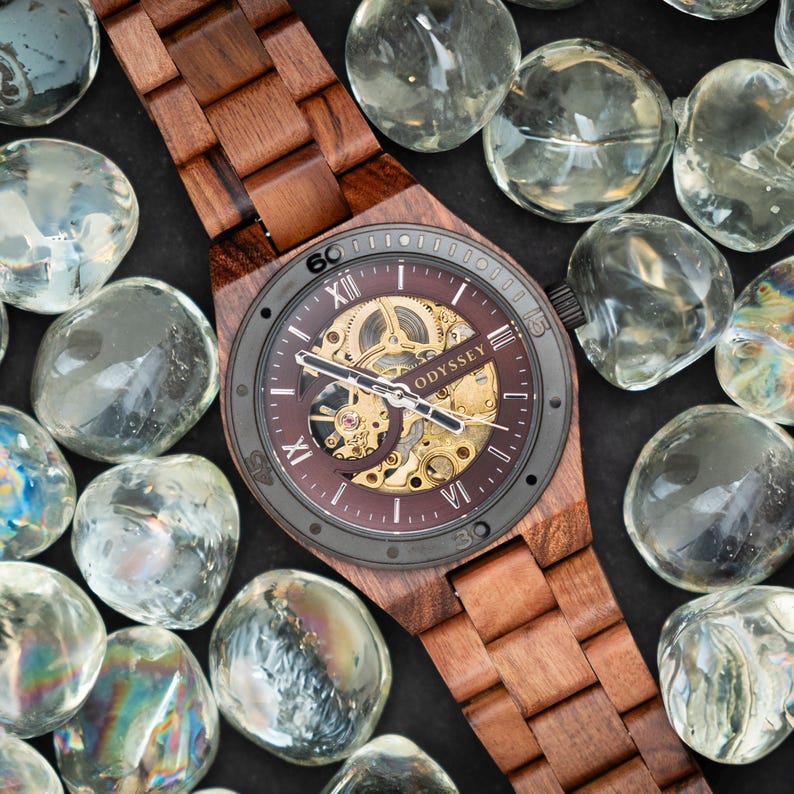 May include: A brown wooden wristwatch with a black face and a skeletonised automatic movement. The watch face has Roman numeral hour markers and the word "ODYSSEY" printed on it.