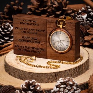 Pode incluir: Um suporte de madeira para relógio de bolso com um relógio dourado de mostrador prateado e algarismos romanos. O bloco de madeira tem gravado o texto: CUSTOM ENGRAVING, TEXT IN ANY FONT OR STYLE, YOUR OWN HANDWRITING, OR A LINE DRAWING OF YOUR PHOTO.