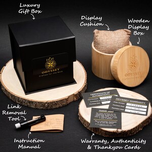 May include: A black gift box with a gold logo that reads "ODYSSEY Crafted for the Bold" sits on a wooden base. The box is surrounded by other packaging materials, including a wooden display box, a display cushion, a link removal tool, an instruction manual, a certificate of authenticity, a warranty card, and a thank you card.