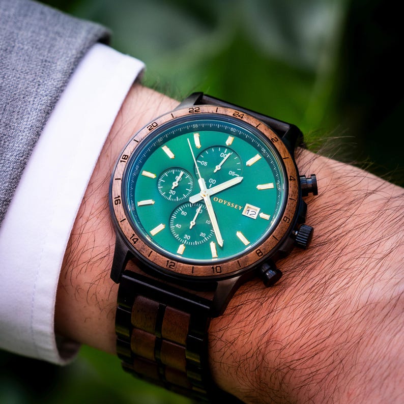 A beautiful wooden watch, with green face surrounded by a walnut bezel accompanied by a walnut and black stainless steel strap. On a mans wrist next to a white and grey suit.