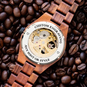 May include: A wooden wristwatch with a brown wood band and a silver face. The back of the watch is open, revealing the inner workings of the watch. The watch face has the text "CUSTOM ENGRAVING ANY FONT OR STYLE" around the edge.
