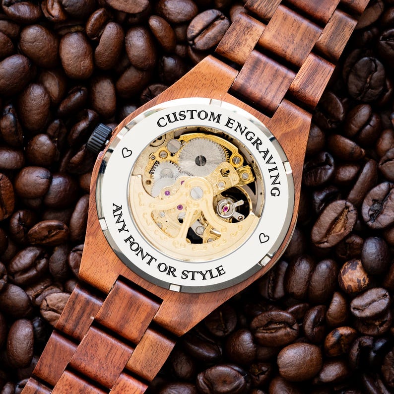 May include: A wooden wristwatch with a brown wood band and a silver face. The back of the watch is open, revealing the inner workings of the watch. The watch face has the text "CUSTOM ENGRAVING ANY FONT OR STYLE" around the edge.