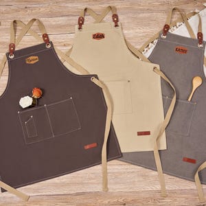 Puede incluir: Tres delantales de lona en marrón, beige y gris, cada uno con un bolsillo delantero y detalles de cuero. Los delantales tienen correas ajustables y están personalizados con los nombres: "Barton", "Edith" y "Kathy". Una cuchara de madera y flores están en los bolsillos.