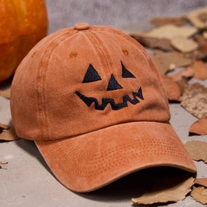 Puede incluir: Gorra de béisbol naranja con un diseño de cara de calabaza negra. La gorra tiene una visera curva y está hecha de una tela suave y texturizada. Perfecta para Halloween o festividades de otoño.