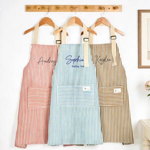 Puede incluir: Tres delantales a rayas en coral, turquesa y marr&oacute;n cuelgan de perchas de madera. Cada delantal tiene un bolsillo delantero grande y est&aacute; personalizado con un nombre en escritura cursiva. El delantal del medio dice "Sophia Baking Star".