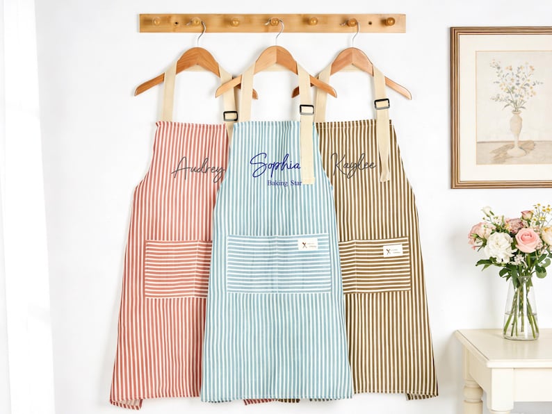 Puede incluir: Tres delantales a rayas en coral, turquesa y marr&oacute;n cuelgan de perchas de madera. Cada delantal tiene un bolsillo delantero grande y est&aacute; personalizado con un nombre en escritura cursiva. El delantal del medio dice "Sophia Baking Star".