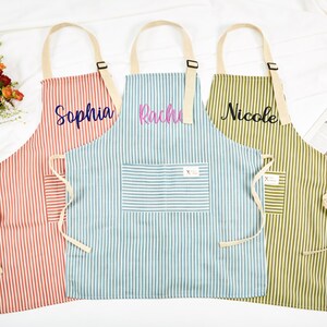 Puede incluir: Tres delantales personalizados con rayas verticales en coral, azul y verde oliva. Cada delantal tiene un nombre bordado: Sophia, Rachi y Nicole. Los delantales tienen correas y bolsillos color crema.