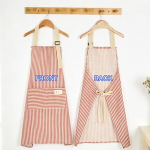 Puede incluir: Dos delantales a rayas rojas y blancas colgando de perchas de madera. El delantal delantero tiene un bolsillo y la palabra "FRONT" en azul. El delantal trasero est&aacute; atado a la cintura y tiene la palabra "BACK" en azul. Un jarr&oacute;n de flores est&aacute; sobre una mesa.