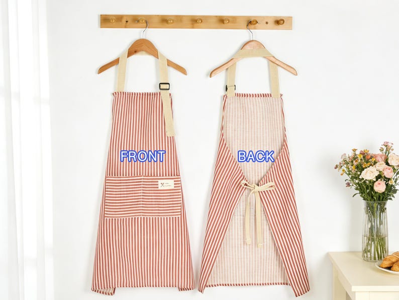 Puede incluir: Dos delantales a rayas rojas y blancas colgando de perchas de madera. El delantal delantero tiene un bolsillo y la palabra "FRONT" en azul. El delantal trasero est&aacute; atado a la cintura y tiene la palabra "BACK" en azul. Un jarr&oacute;n de flores est&aacute; sobre una mesa.
