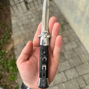 Puede incluir: Una navaja plegable plateada y negra con una hoja puntiaguda. El mango es negro con un patrón veteado. El cuchillo se sostiene en una mano, con una correa de reloj negra visible.