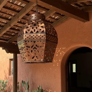 May include: A hanging pendant light fixture with a hexagonal, copper-toned metal frame featuring intricate, laser-cut geometric patterns. The lantern-style shade has a weathered, textured finish and is suspended by a metal chain from a wooden beam.