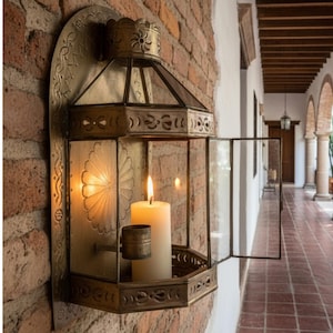 May include: A wall-mounted brass lantern with a hexagonal frame, glass panels, and intricate cut-out patterns. The lantern features a hinged door and a central brass candle holder containing a single lit white pillar candle, casting a warm glow.