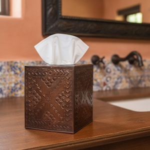 May include: A decorative brown tissue box with an embossed floral and arrow design. White tissues are visible at the top. The box is made of metal and sits on a wooden surface. The bathroom decor includes a mirror and faucet.