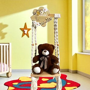 May include: A brown teddy bear sits in a wooden swing with macrame ropes. The swing's top is a flower-shaped wooden sign with the word "world" and floral accents. The swing hangs in a yellow room with a star decoration.