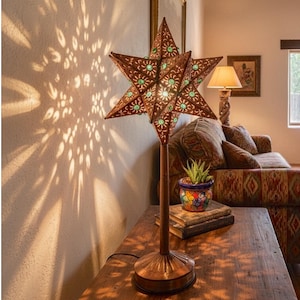 May include: A star-shaped table lamp with a metallic copper finish and green glass accents. The lamp features intricate cutouts that cast a floral light pattern onto the wall. It stands on a circular base with a slender stem supporting the star shade.