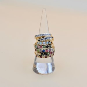 May include: A stack of gold rings with colourful gemstones, including blue, green, red, and white. The rings are displayed on a clear cone-shaped stand.