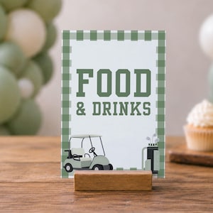 Op de afbeelding: Een rechthoekig bord met een groen-witte gingham rand, voorzien van de tekst FOOD & DRINKS in een donkergroen bloklettertype. Het bord toont een kleine illustratie van een golfkarretje en een golftas, geplaatst in een houten blokstandaard.