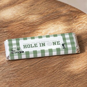 Pode incluir: Uma embalagem retangular de barra de chocolate com um padrão de xadrez vichy verde e branco. O centro exibe o texto 'THANK YOU FOR MAKING TODAY A HOLE IN ONE' com um gráfico de bola de golfe, carrinho de golfe e saco de golfe.