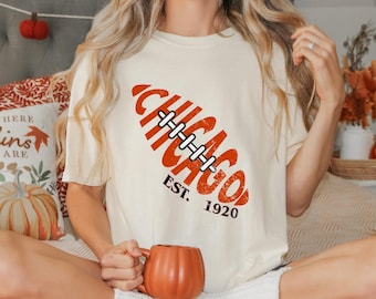 May include: Cream-colored t-shirt featuring a football design with the word "CHICAGO" in orange, along with "EST. 1920". The shirt is a casual style, suitable for everyday wear. A person is holding a pumpkin-shaped mug.