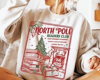 Puede incluir: Una sudadera blanca con un diseño gráfico rojo y verde que presenta una chimenea, una pila de libros, un árbol de Navidad y el texto "North Pole Readers Club" con el lema "Every Wednesday in December, Naughty & Nice List Welcomed, Sleighing The Lists".