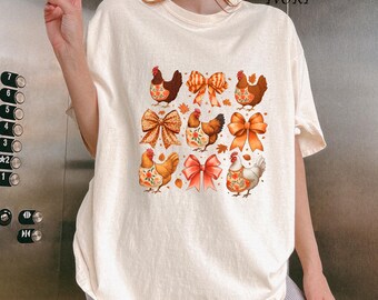 May include: Cream-colored t-shirt featuring a grid of chickens and bows in autumnal colors. The chickens wear floral aprons, and the bows are in shades of orange and red. The word "IVORY" is printed at the top right.