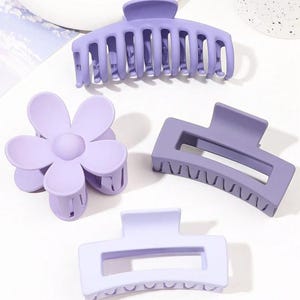 May include: Four lavender hair claw clips in various shapes: a large claw, a flower, and two rectangular clips. The clips are made of a matte plastic material and are arranged on a white surface.