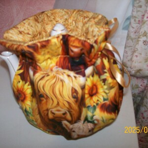 Puede incluir: Una funda para tetera de color marrón y amarillo con un diseño de una vaca de las Highlands y girasoles. La funda está hecha de tela y tiene un cierre de cordón.