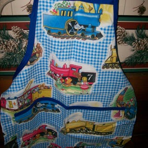 May include: Blue and white gingham apron with a blue trim and straps. The apron features colorful illustrations of trains, including a blue steam engine and a red train with the number 7. The date 2026/04/28 is printed on the bottom.