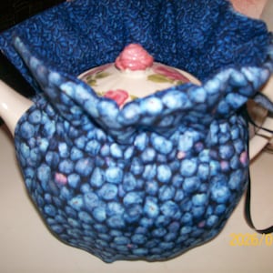 May include: A white ceramic teapot with a floral design, covered by a blue fabric tea cozy. The cozy has a pattern of blue and purple shapes. The lid of the teapot has a pink knob.