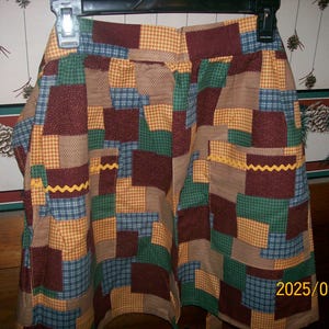 Puede incluir: Falda patchwork con cuadrados de tela en tonos marrón, verde, azul y amarillo. La falda tiene una cintura y un ribete amarillo en zigzag. La fecha "2025/09/29" está impresa en la falda. Cuelga de una percha negra.